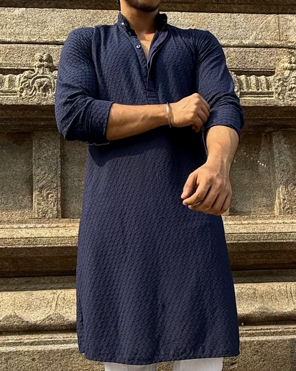 Man in a navy kurta and white pants standing in front of stone architectural structures.
