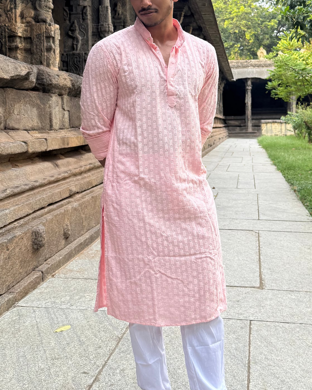 Man in a pink kurta standing in front of an ancient temple structure with greenery in the background.