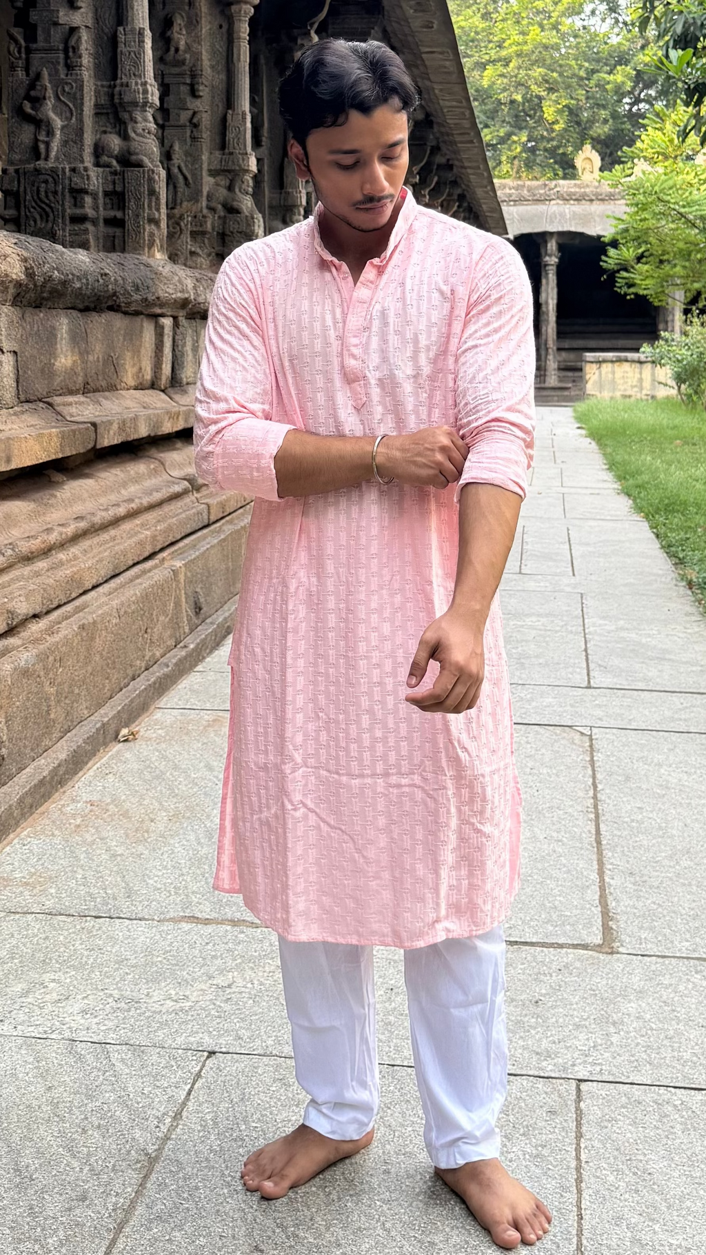 Man in a pink kurta standing on a stone pathway with greenery and an ancient structure in the background